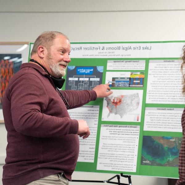 A person points to a spot on a board containing information on research on Lake Erie algal blooms while talking with others.