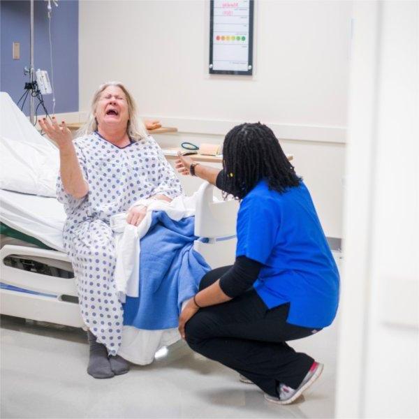nursing student in blue scrub top kneels before a standardized patient who is in a hospital bed